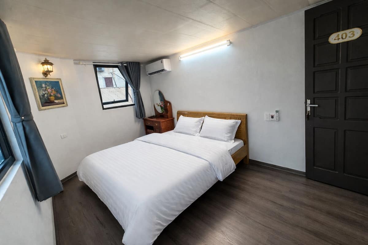 Quarto de hotel com cama, penteadeira, espelho, janela com cortinas, ar condicionado e porta com placa de número.