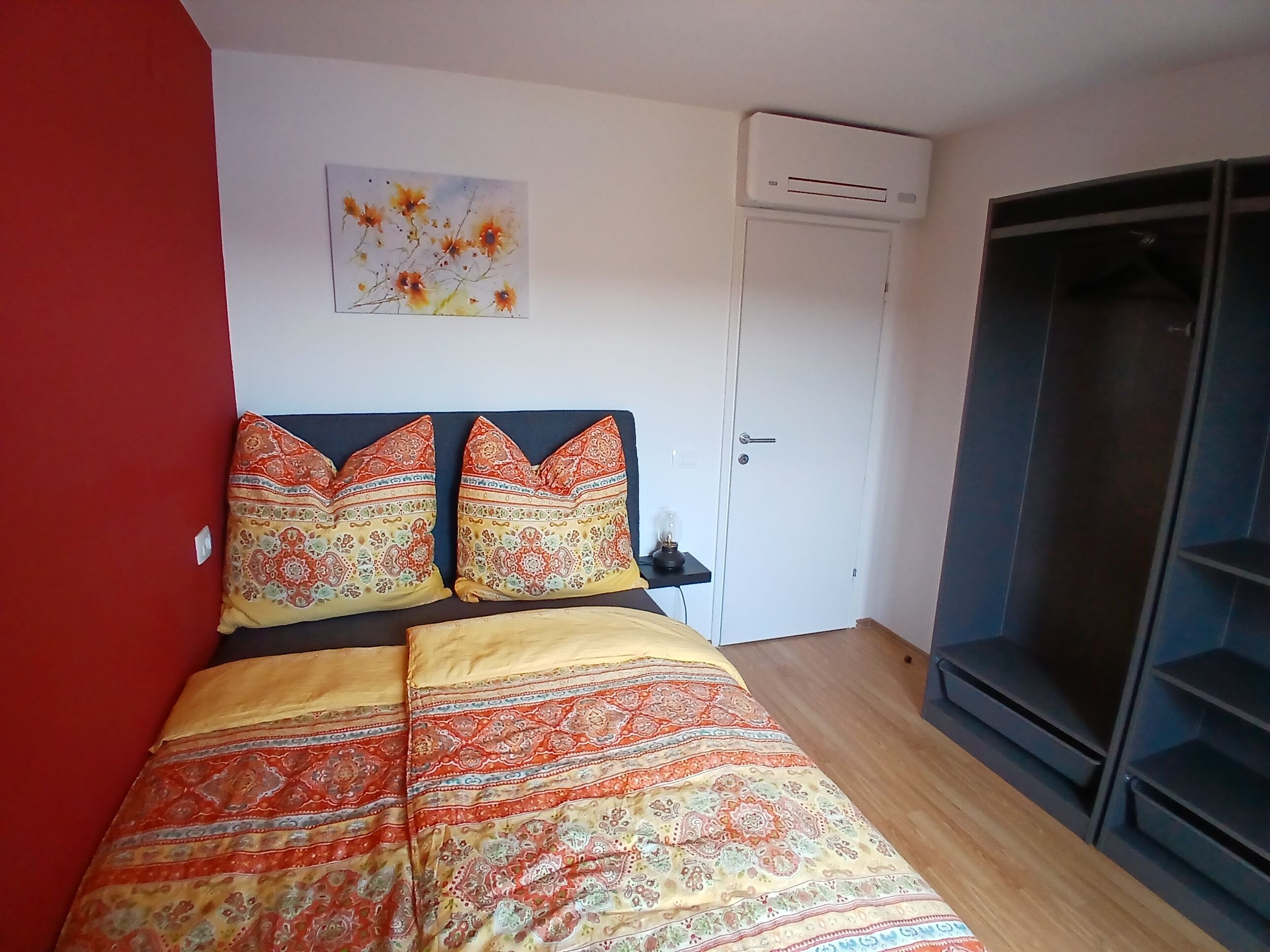 A bedroom with a bed, a red wall, a white door, and a window.