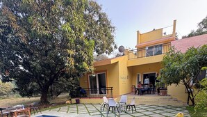 A swimming pool with a tiled deck, surrounded by a garden and a building with a yellow facade. - 4BHK Pool Villa In Alibaug- Anushashi's Villa (Alibag)