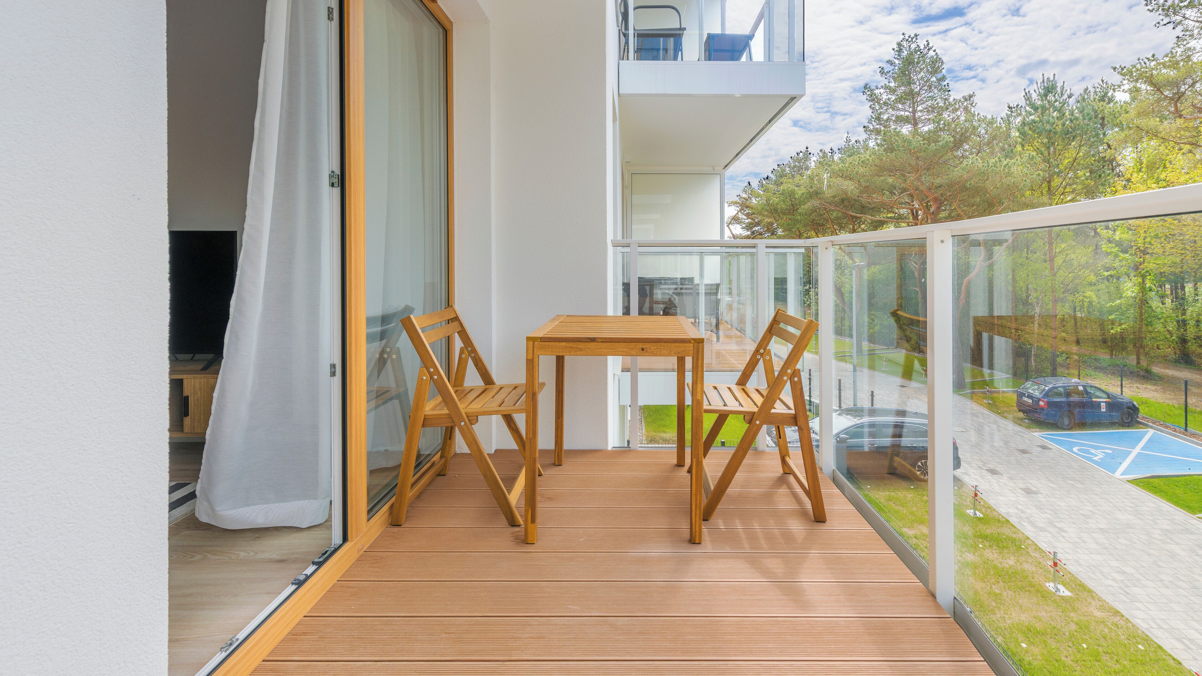 A balcony with a wooden table and chairs, a glass railing, and a view of a parking area and trees.