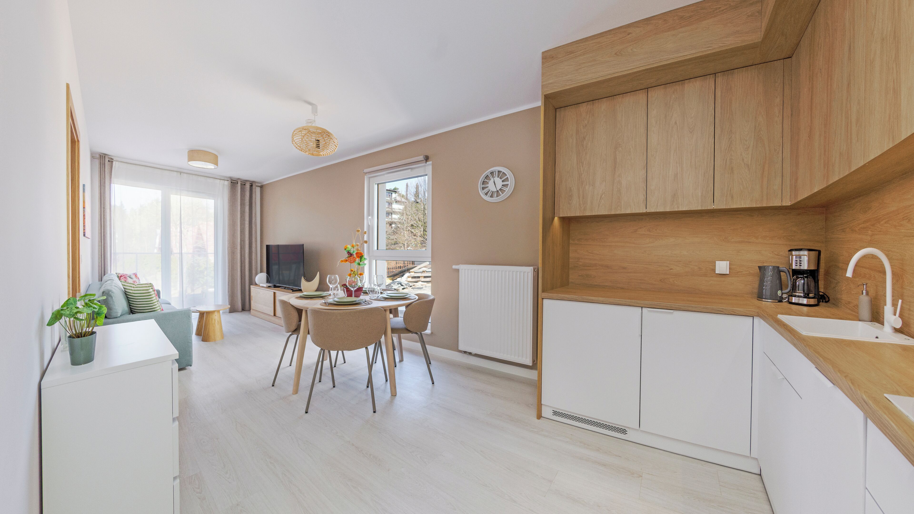 Une cuisine moderne avec des meubles en bois, un coin repas avec une table dressée pour quatre personnes, et un espace de vie avec un canapé et un téléviseur.