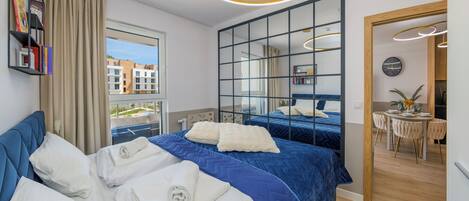 A modern hotel room with a large bed, a view of buildings, a dining area, and a bathroom.