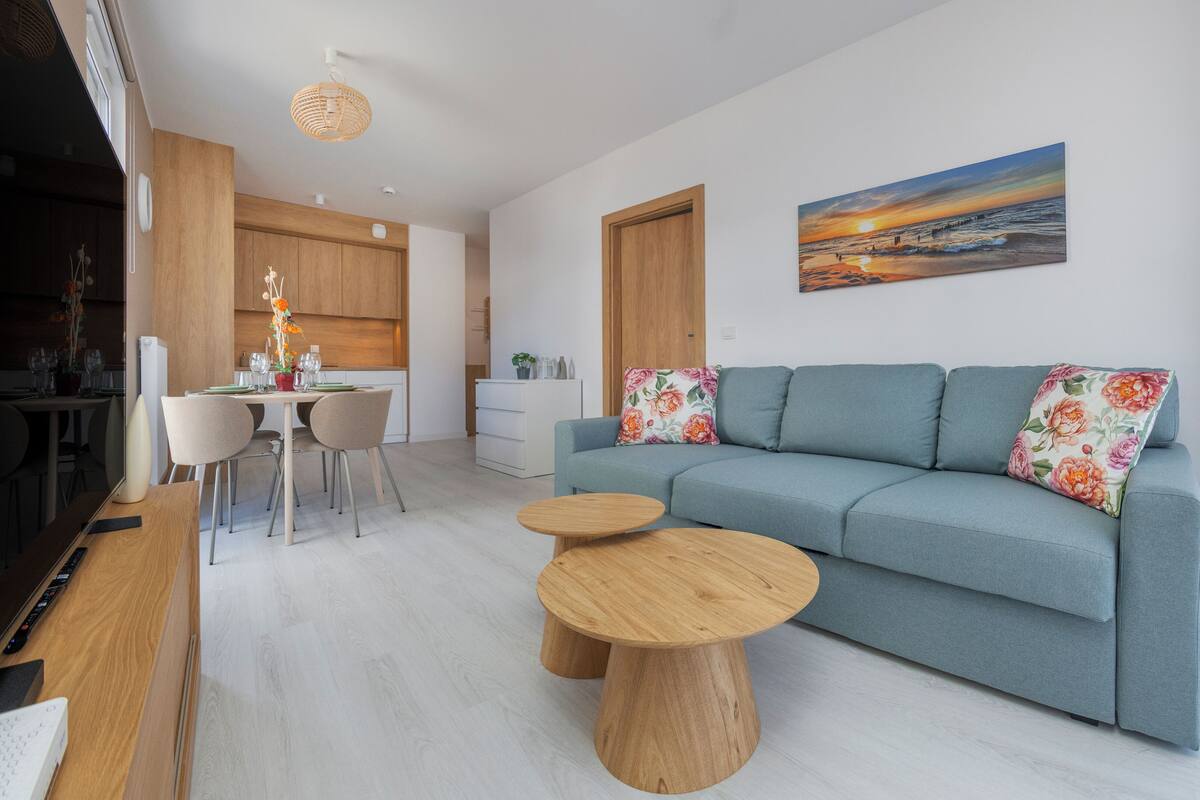 Un salon moderne avec un canapé, deux petites tables rondes en bois, un coin repas avec des chaises et, au fond, une cuisine.