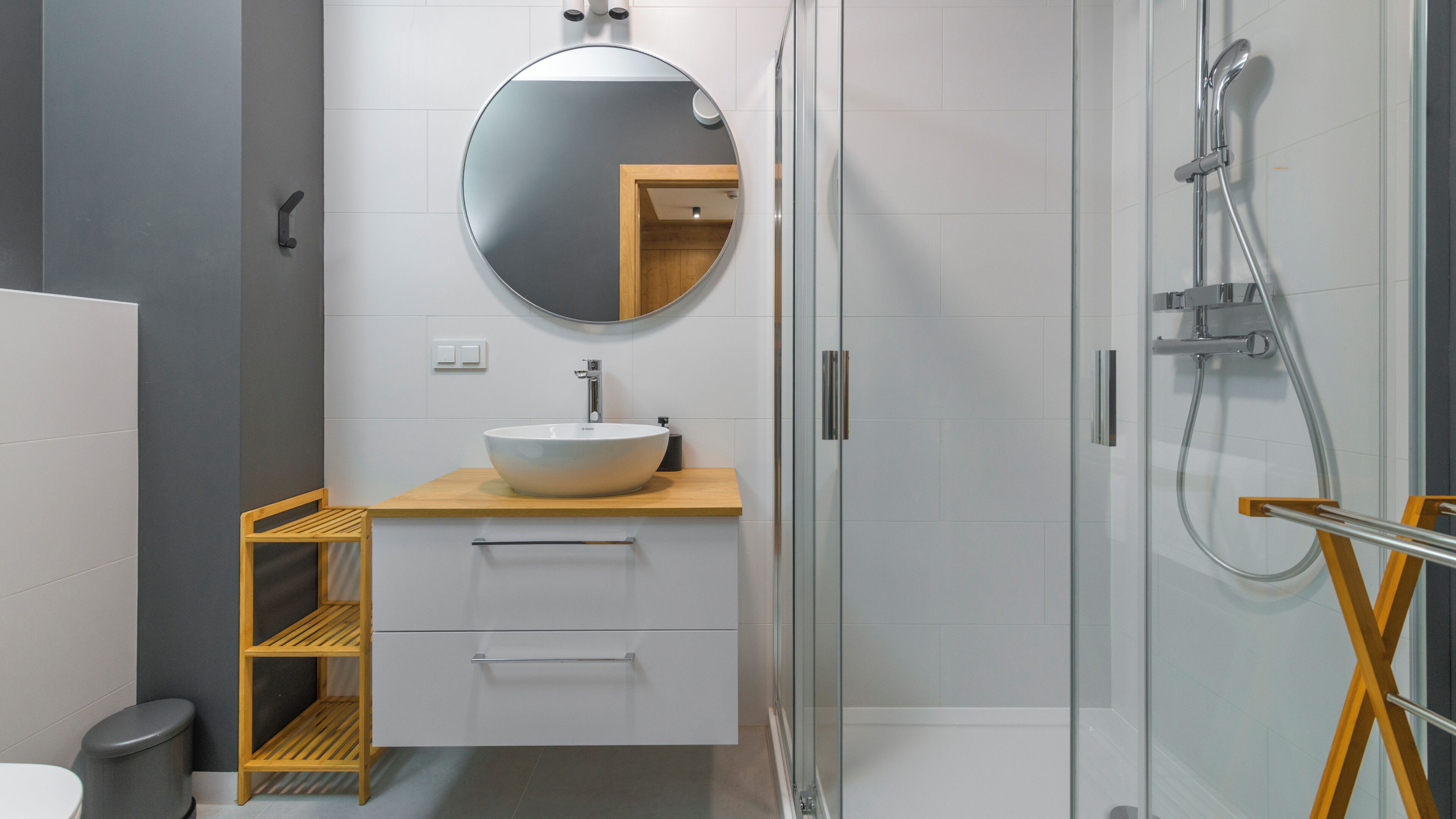 A modern bathroom with a round mirror, a vessel sink, a glass-enclosed shower, and a wooden shelf.