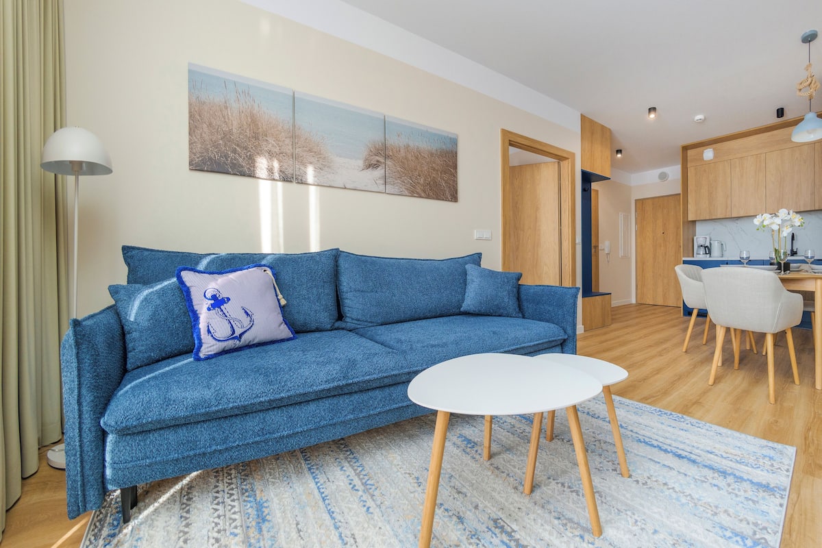Un salon moderne avec un canapé bleu, une petite table blanche et un coin cuisine doté de meubles en bois et d’une table à manger.