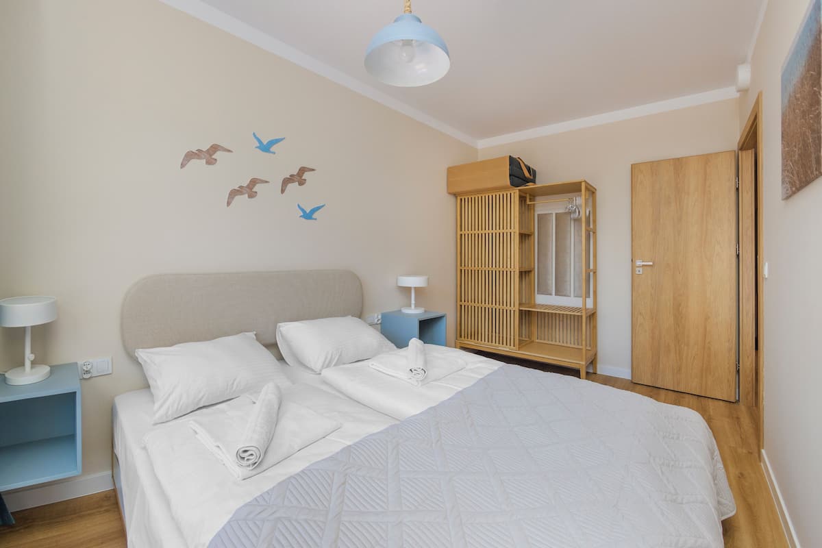 Un lit bien fait, avec du linge de lit blanc, une table de chevet avec une lampe, une armoire et des tableaux représentant des oiseaux accrochés au mur.
