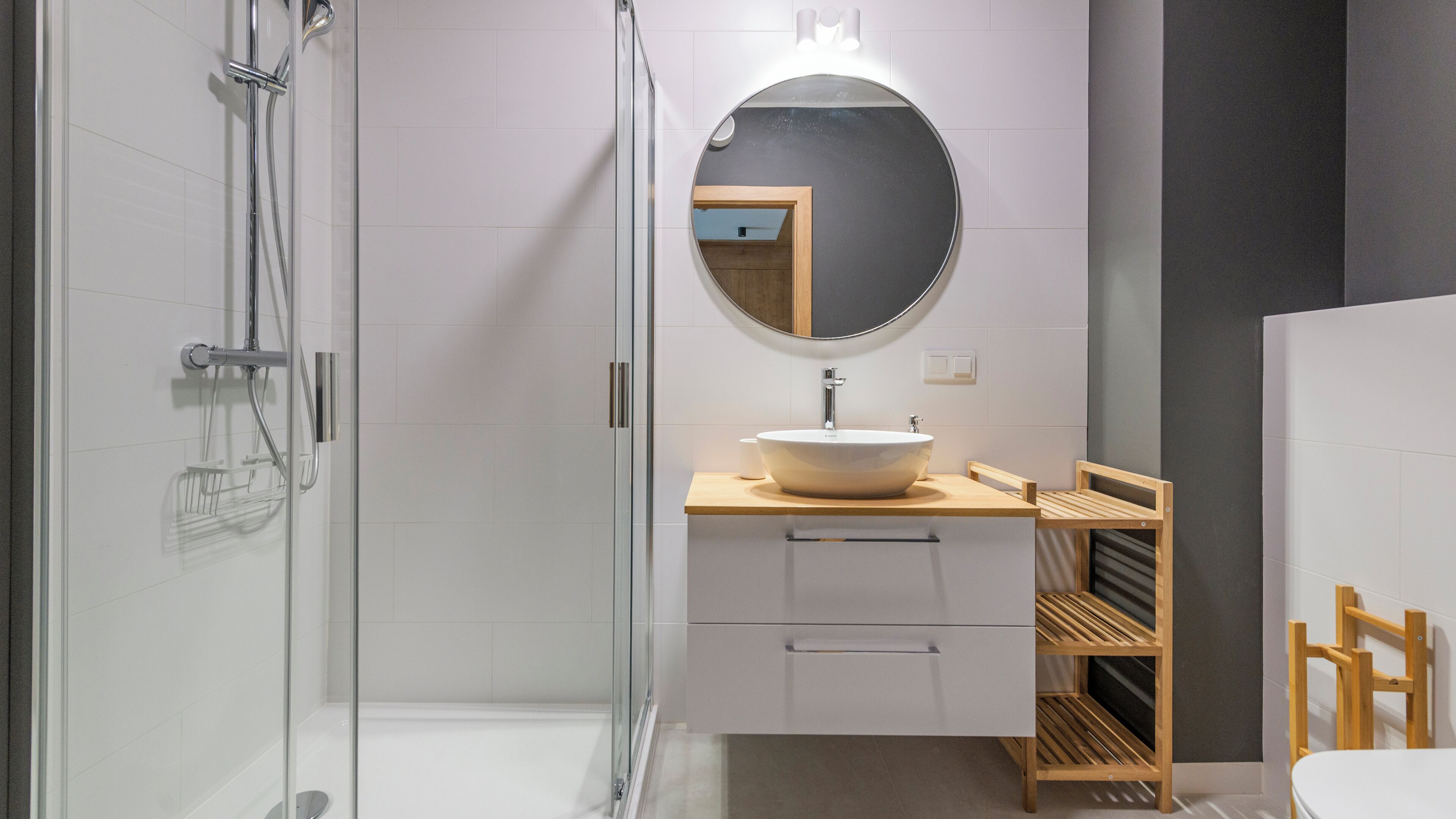 Un baño moderno con cabina de ducha de vidrio, un espejo redondo, un lavabo blanco y estantes de madera.
