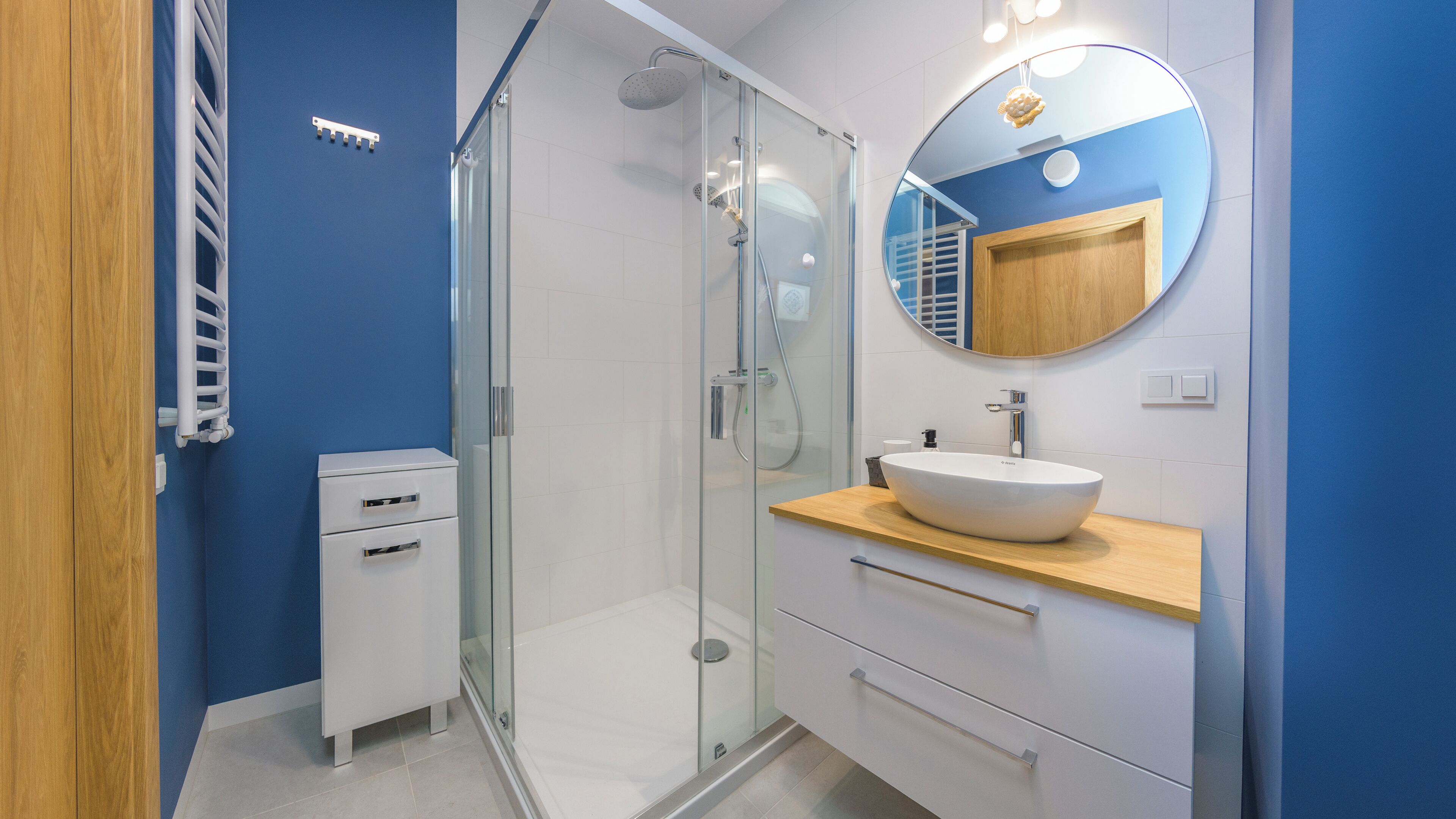 Un baño moderno con cabina de ducha de vidrio, un lavabo blanco con encimera de madera y un espejo redondo sobre el lavabo.
