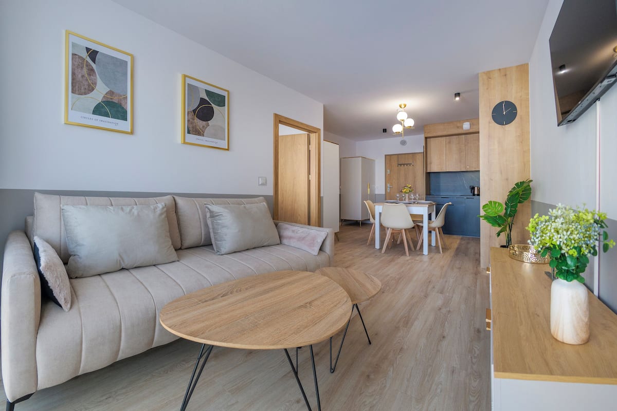Un salon moderne avec un canapé, une table basse ronde en bois et un coin repas comprenant une table et des chaises. Deux œuvres d’art encadrées ornent le mur, on aperçoit une porte en bois et, au fond, un espace cuisine.