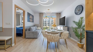 A modern living room with a sofa, a dining table with chairs, a TV, and a large window with a view of buildings. - Apartamenty Sun & Snow Bałtyk Jastrzębia Góra (Jastrzębia Góra)