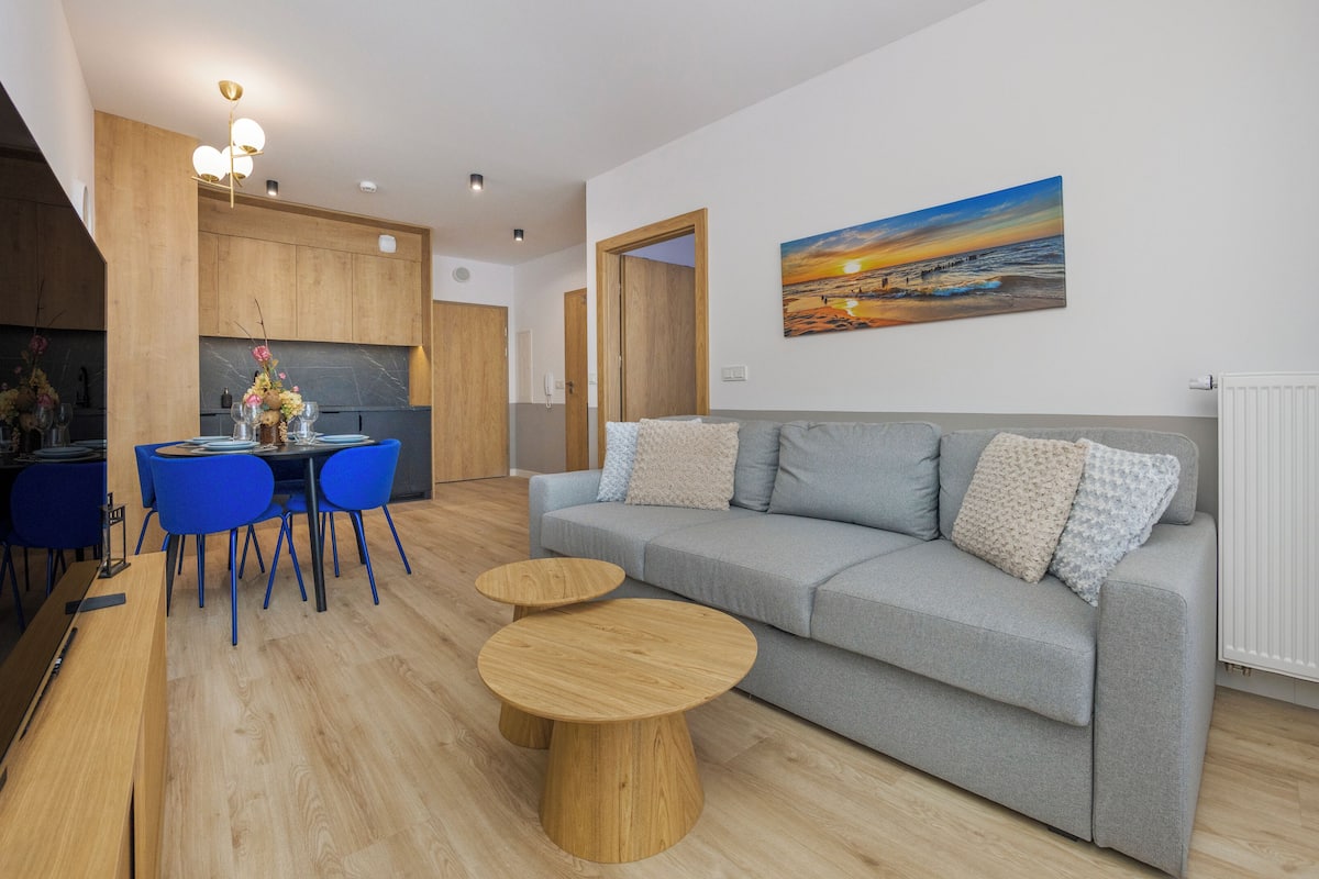 Un salon moderne avec un canapé gris, des fauteuils bleus, une table basse en bois et un tableau représentant un coucher de soleil.