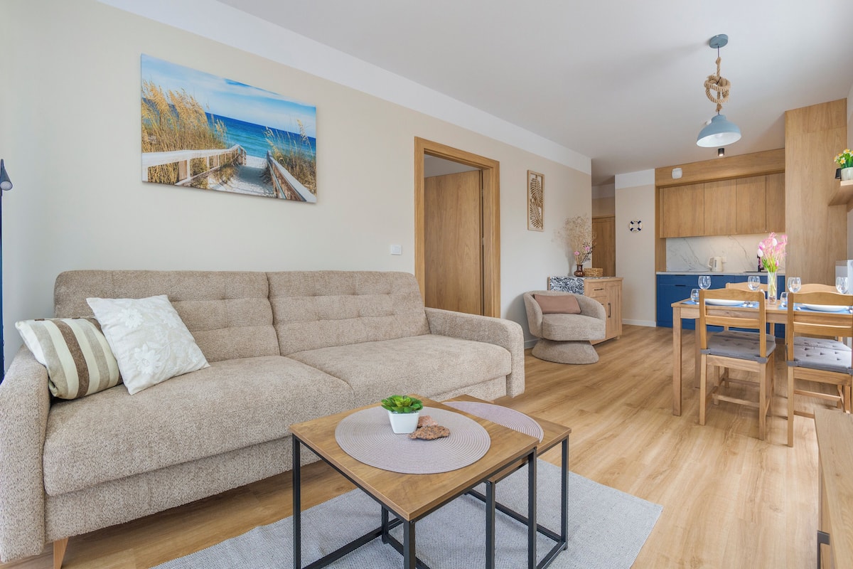 Un salon comprenant un canapé, une table basse, un coin repas avec une table et des chaises, une cuisine avec des meubles en bois et un tableau accroché au mur.