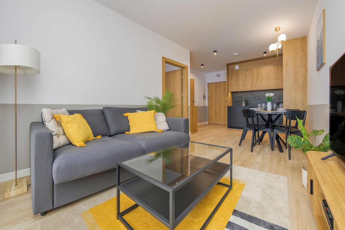 Un salon moderne avec un canapé gris, des coussins jaunes, une table basse en verre et un coin repas comprenant une table et des chaises en bois.