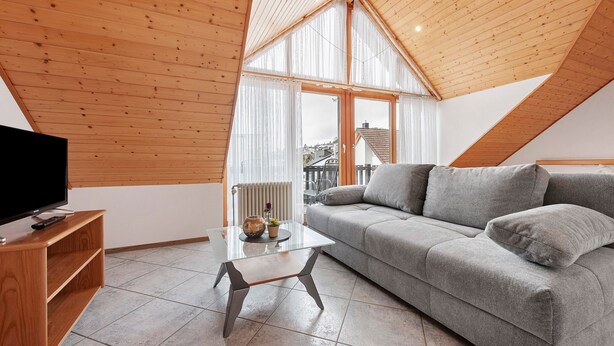 Un soggiorno moderno con soffitto in legno, un divano grigio, un tavolino da caffè in vetro e un televisore su un mobile in legno.