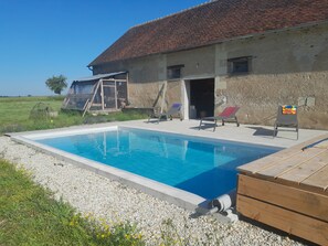 Ein Schwimmbecken mit klarem, blauem Wasser, umgeben von einer Kiesfläche, und im Hintergrund ein Gebäude mit einem Ziegeldach.