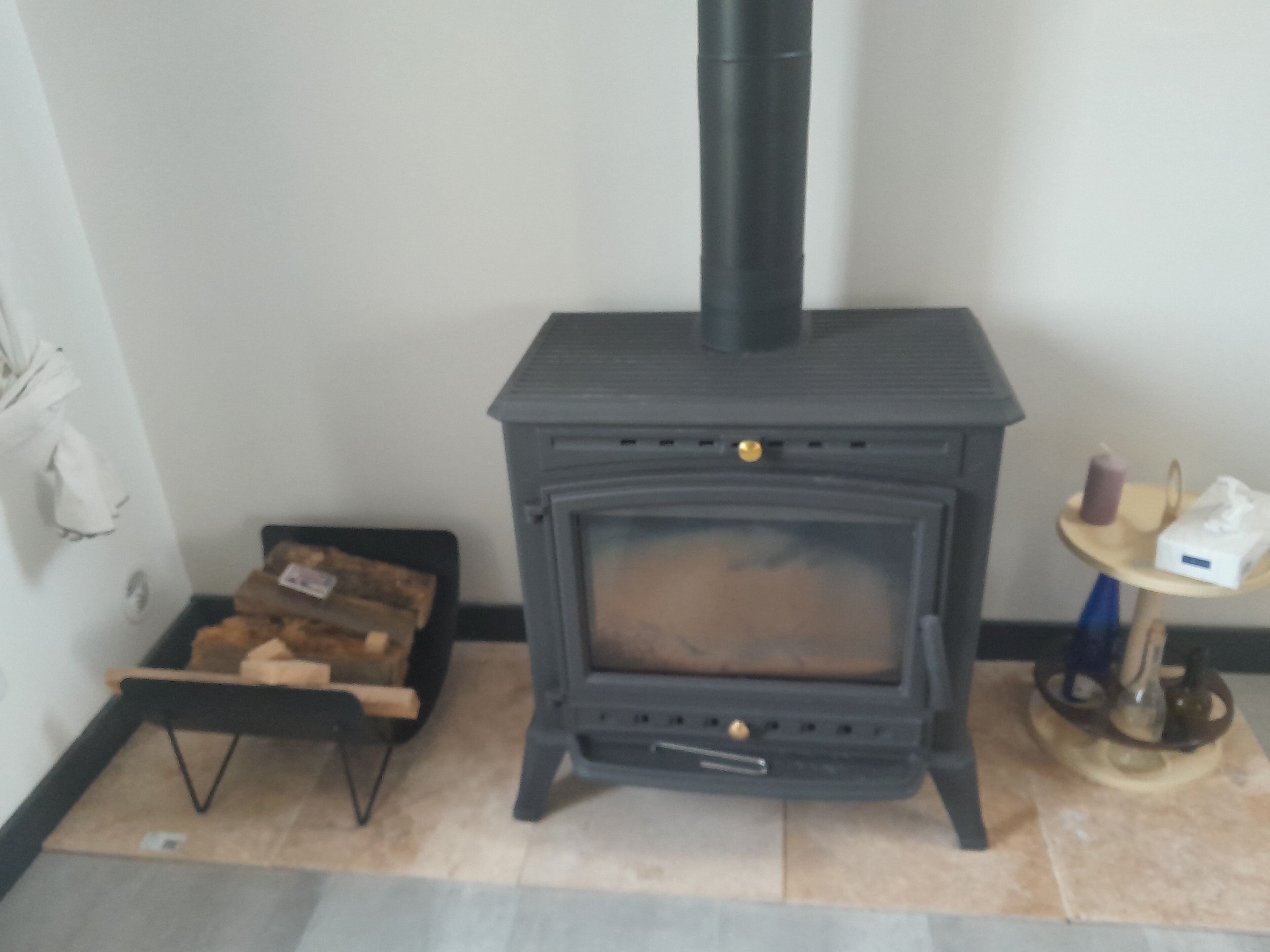 A wood-burning stove with a chimney, a small table with a candle and tissue box, and a box of firewood on the floor.