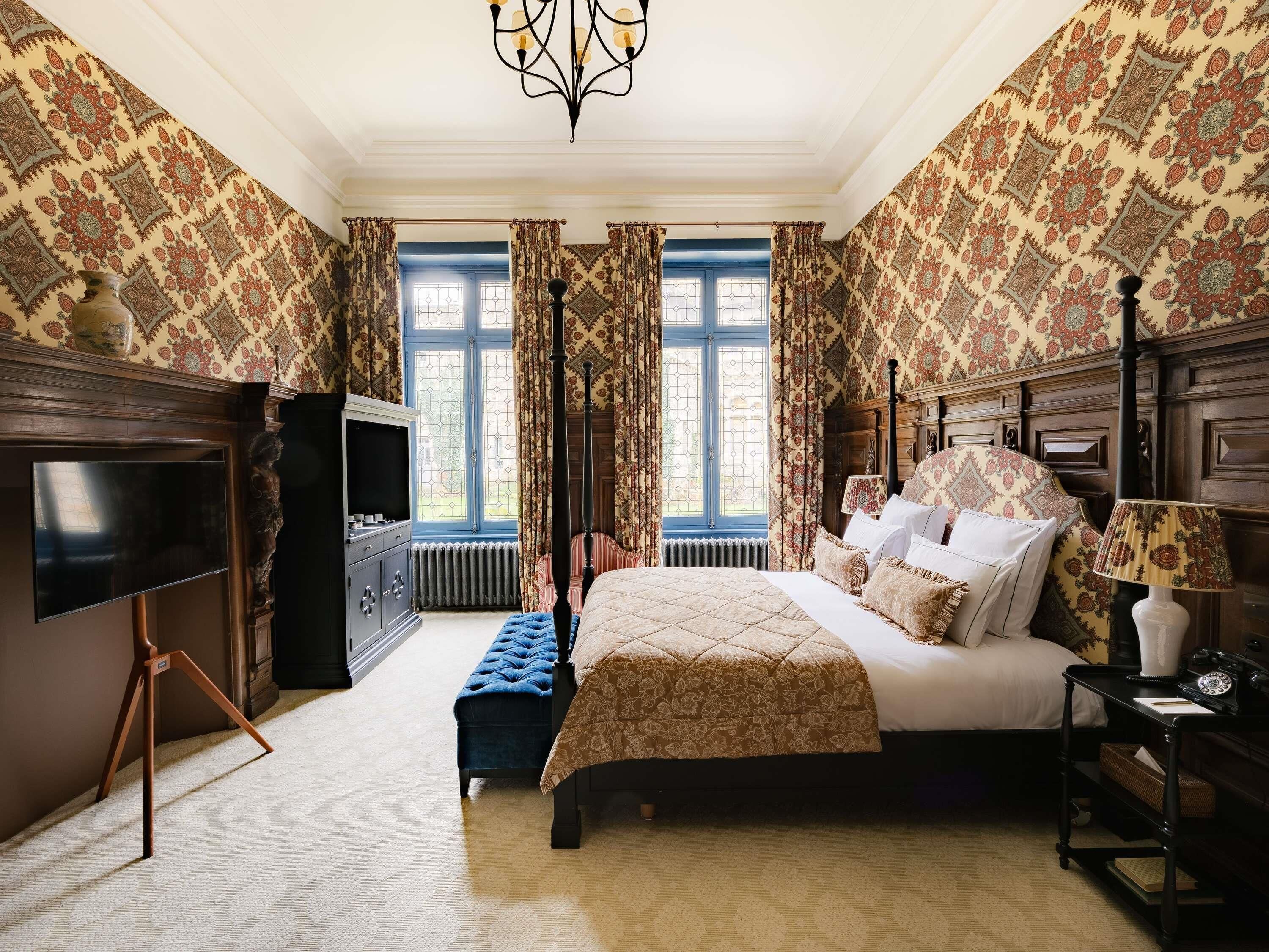 A bedroom with a four-poster bed, a television, a desk with a lamp, and patterned wallpaper.