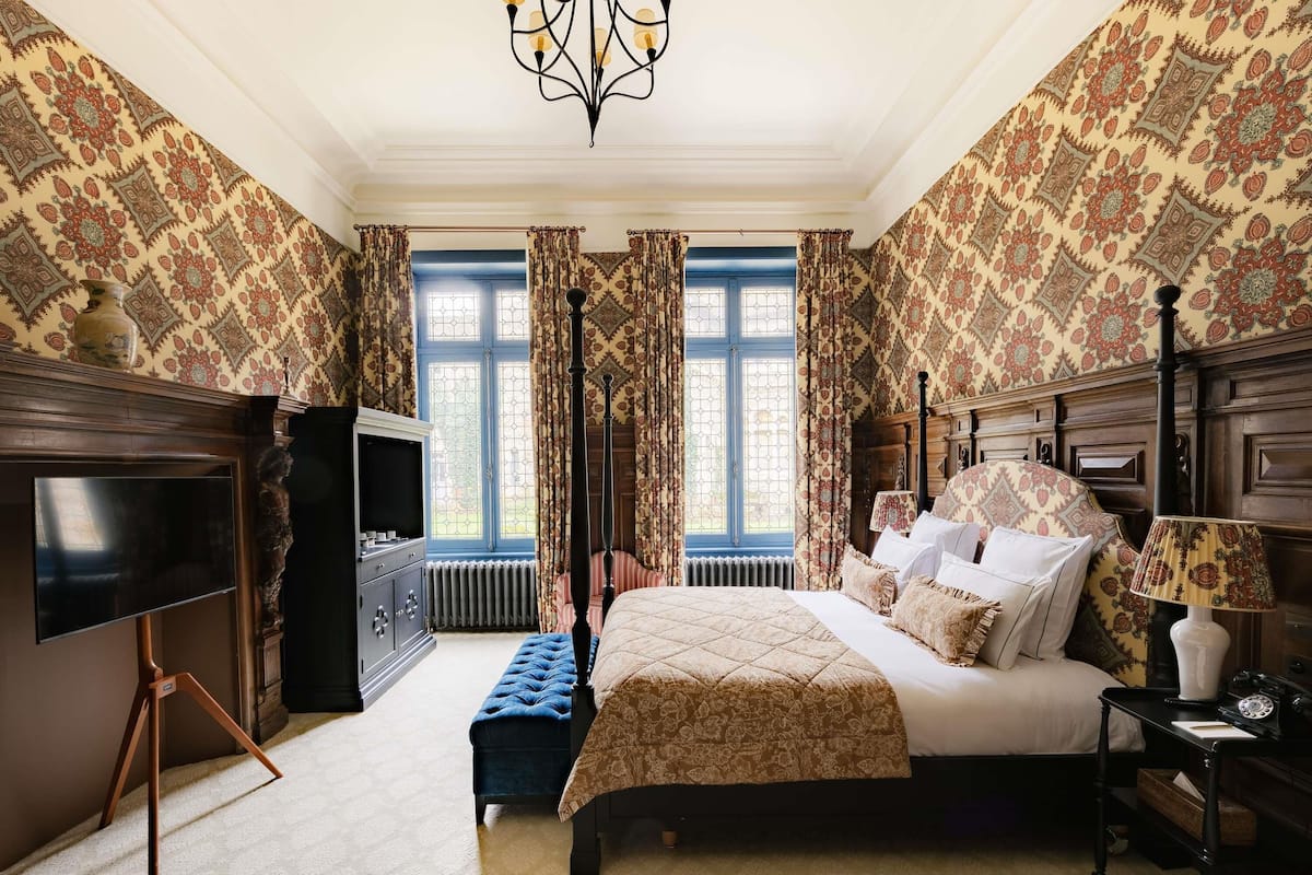 A bedroom with a four-poster bed, a television, a desk with a lamp, and patterned wallpaper.