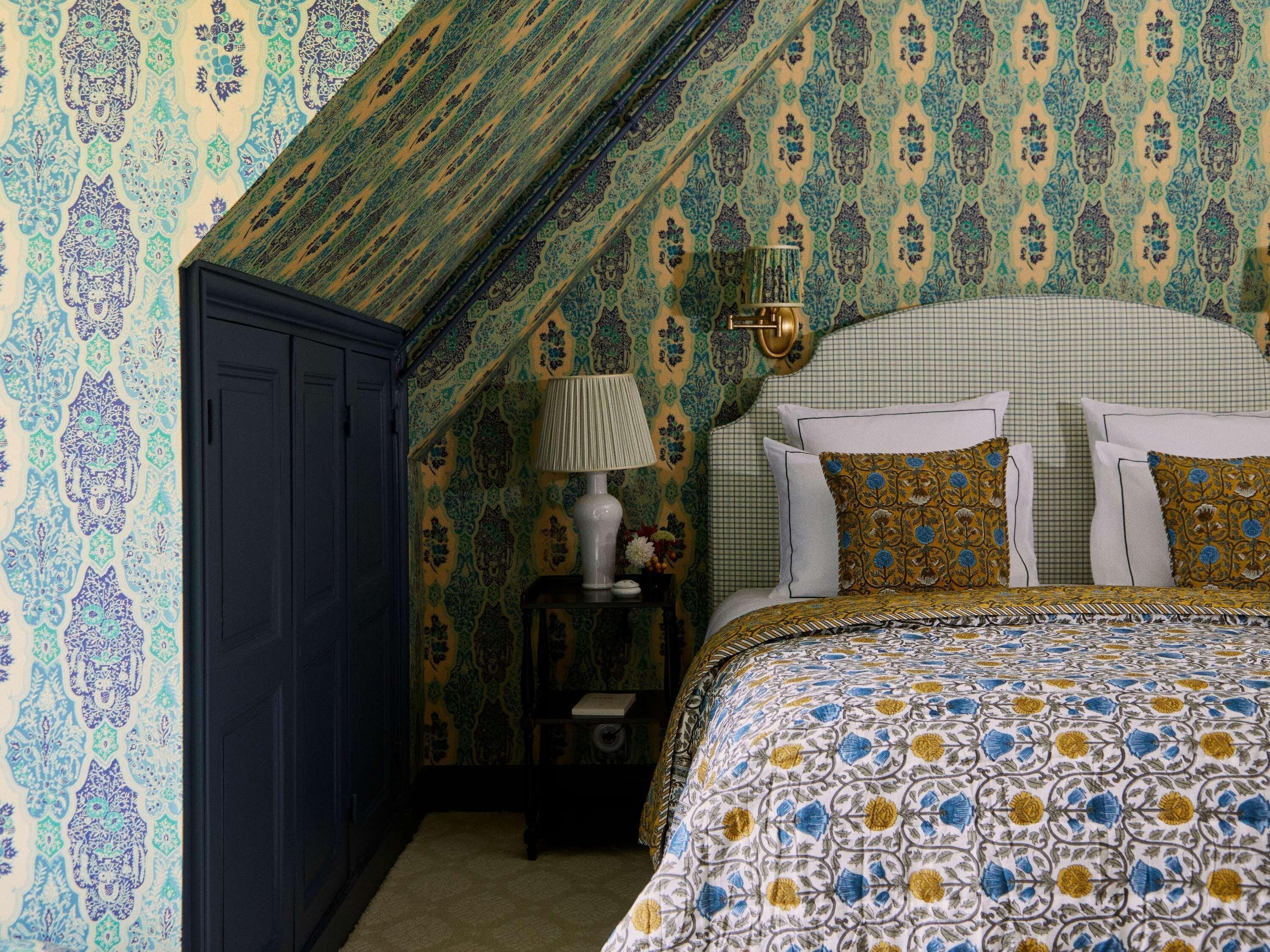 Une chambre avec un plafond en pente, un papier peint à motifs, un lit avec une literie à fleurs, une table de chevet avec une lampe et une armoire sombre.