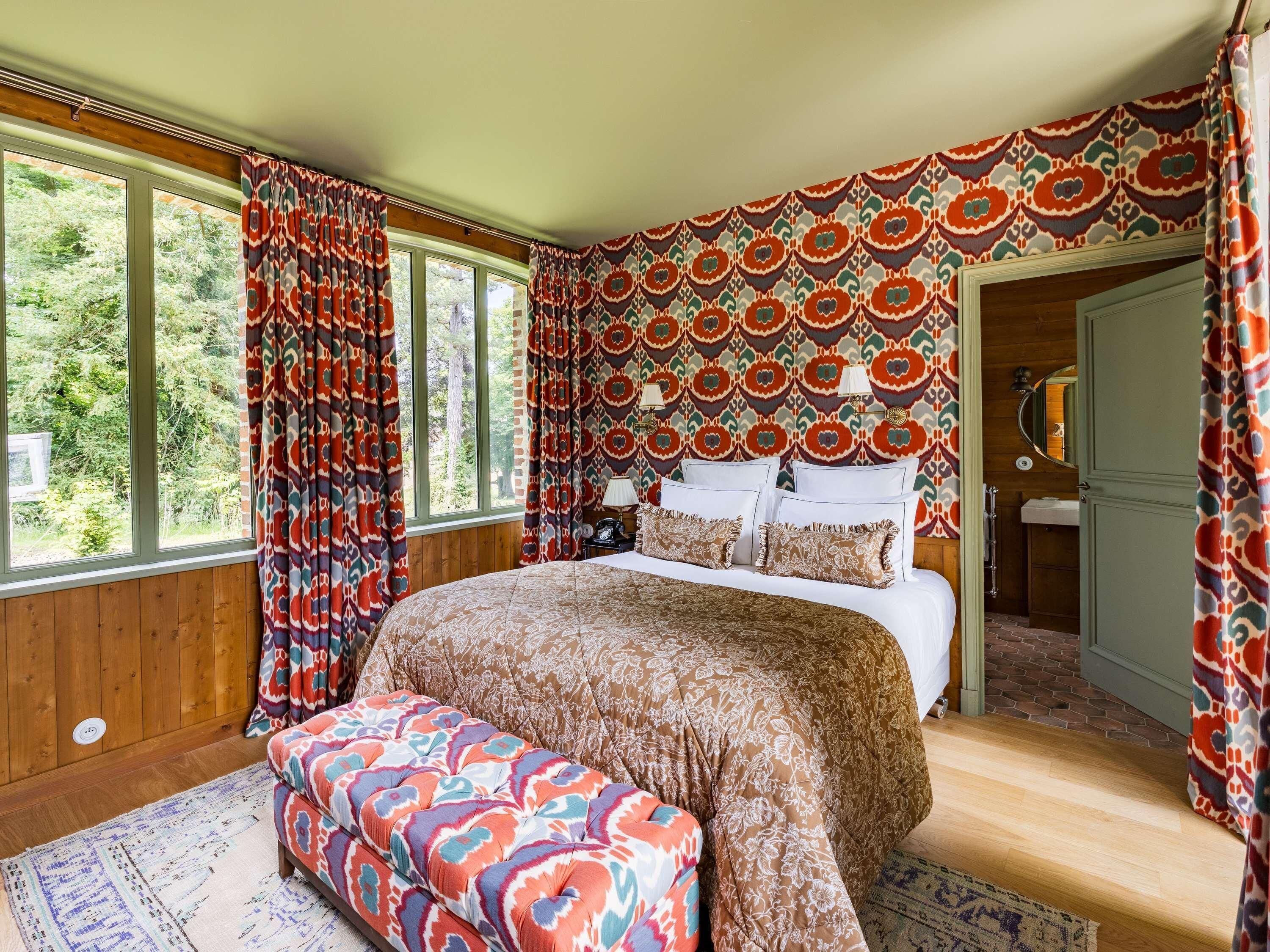 A bedroom with a large bed, a bench, patterned curtains, and a view of the outdoors.