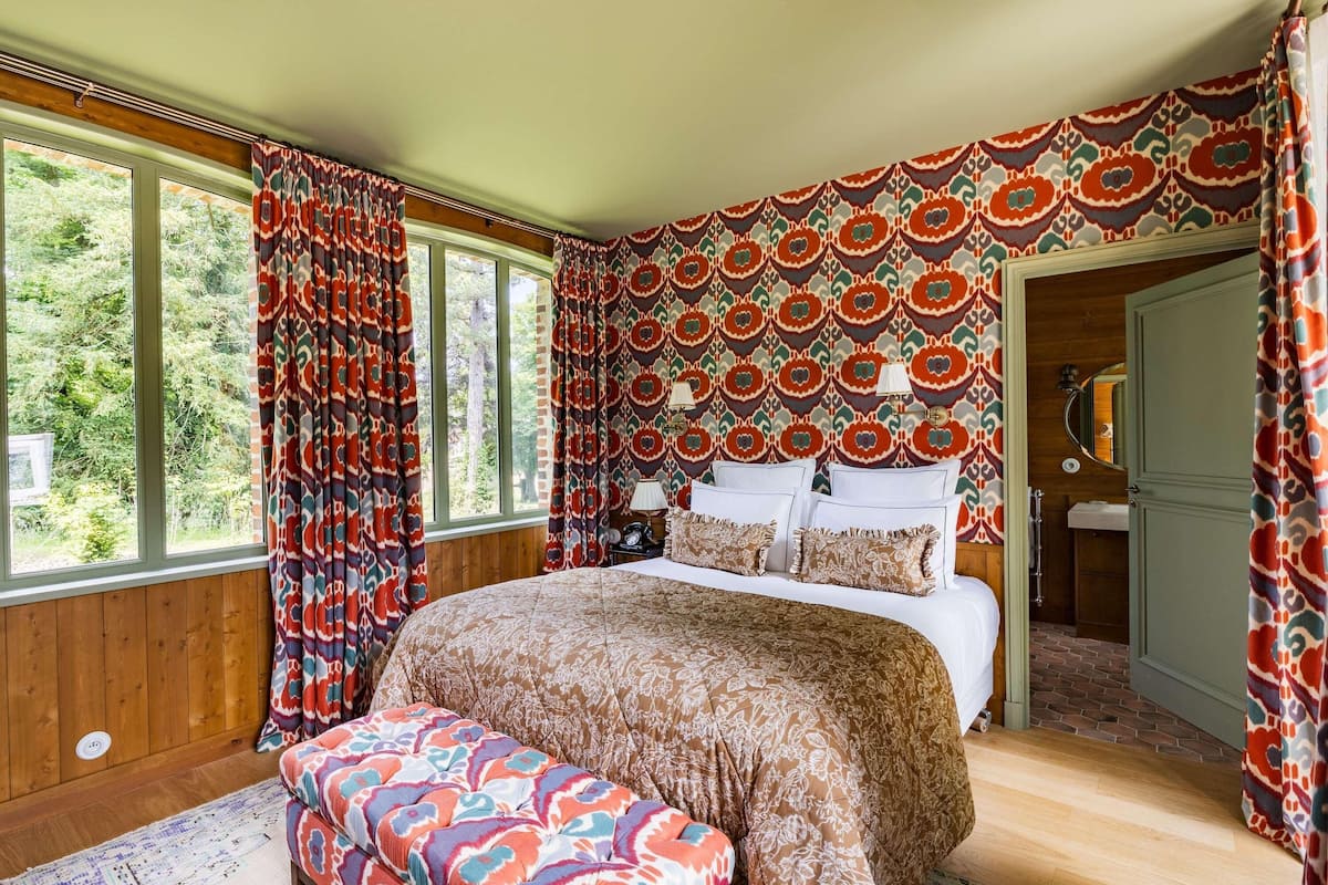 A bedroom with a large bed, a bench, patterned curtains, and a view of the outdoors.