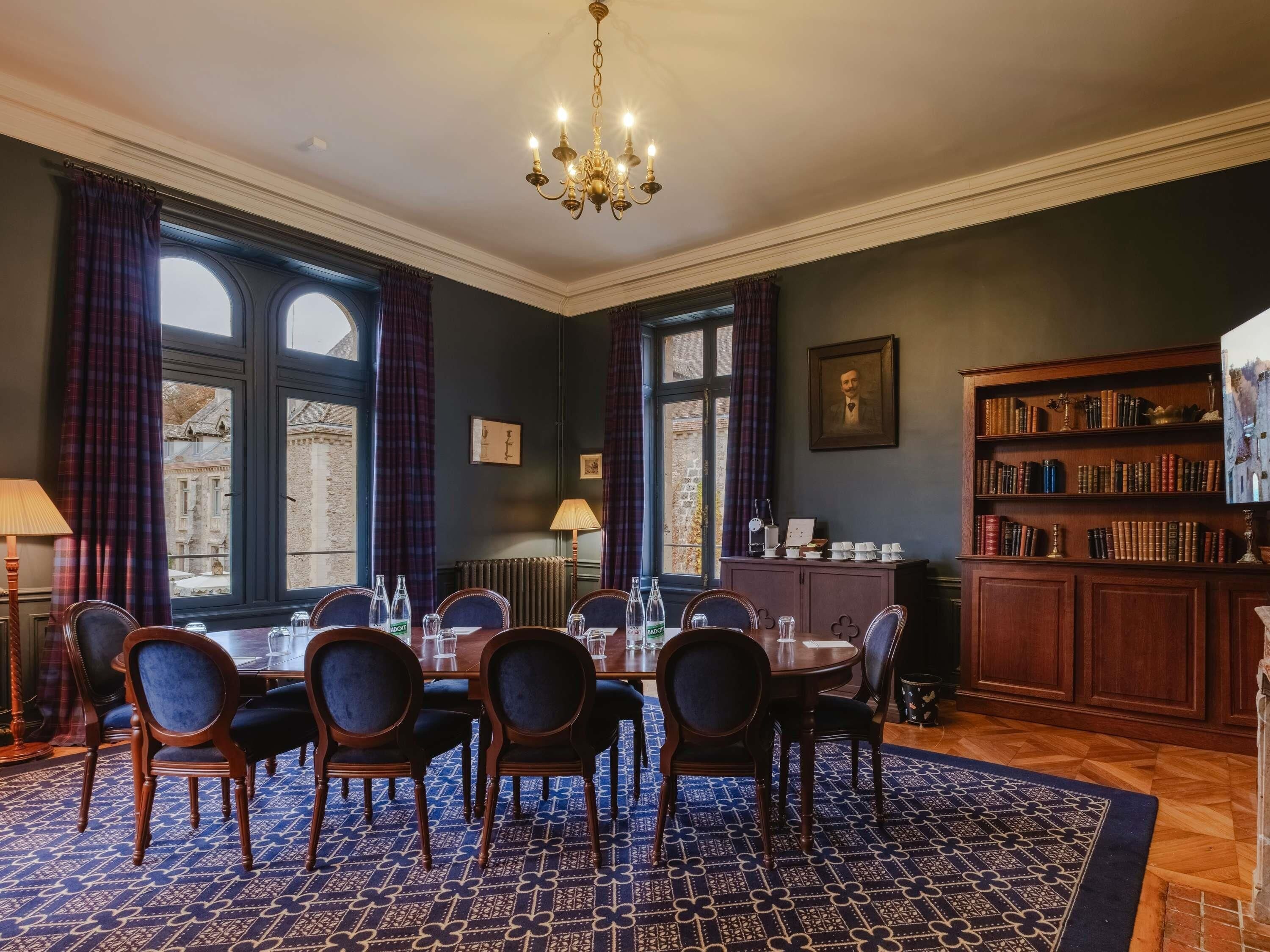 A dining room with a round table, chairs, a chandelier, a bookshelf, and large windows.