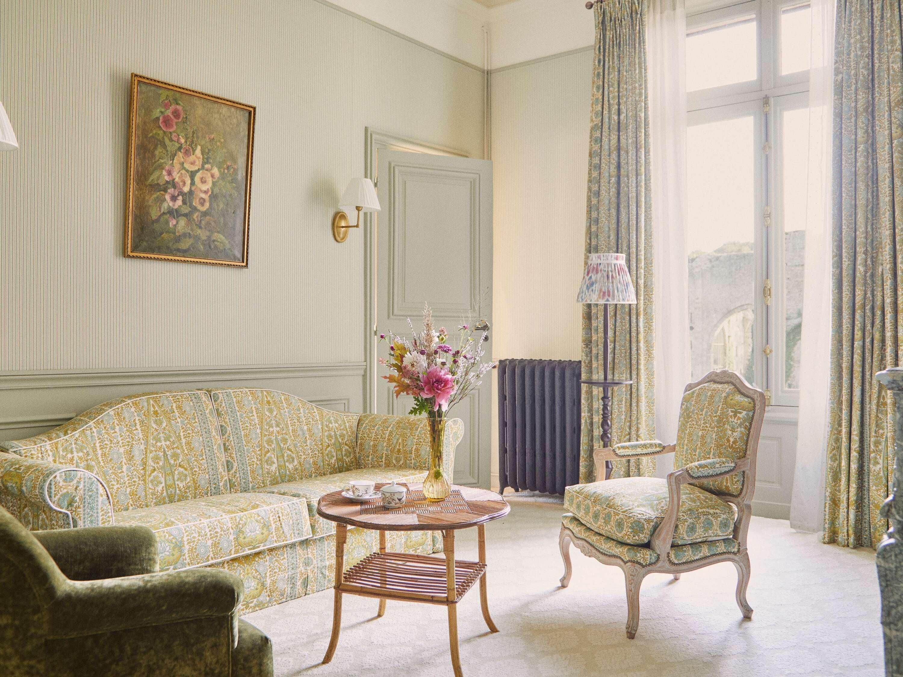 A room with a patterned sofa, a chair, a small table with a vase, a lamp, and a painting on the wall.