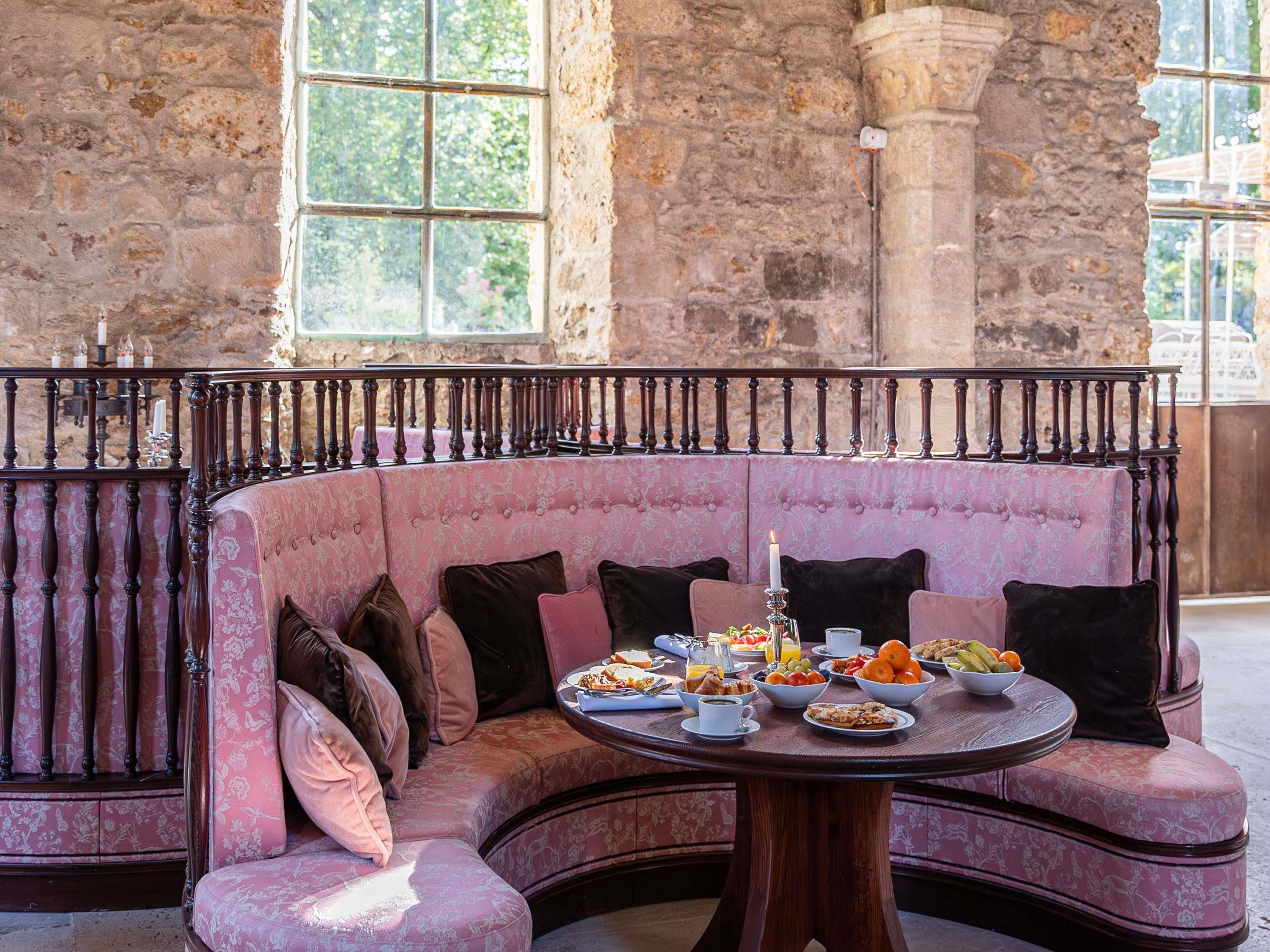 A curved pink sofa with cushions, a round wooden table with breakfast items, and a stone wall with windows.