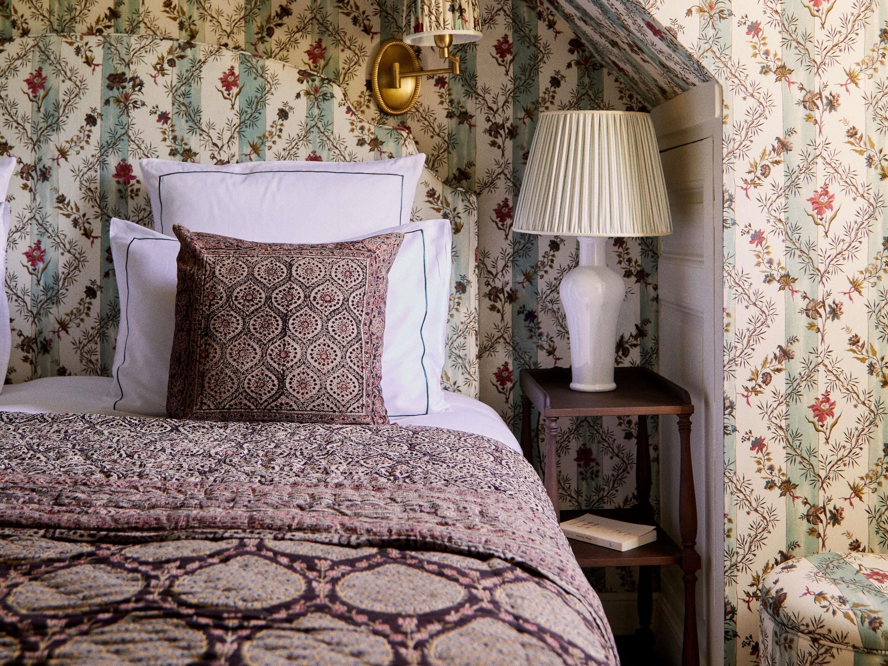 Una camera da letto con una coperta a motivi, cuscini, una lampada da comodino e carta da parati floreale.