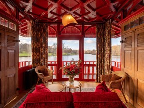 A room with a red interior, a wooden ceiling, a view of a lake, wicker furniture, and a red sofa. - Abbaye Des Vaux De Cernay (Cernay-la-Ville)