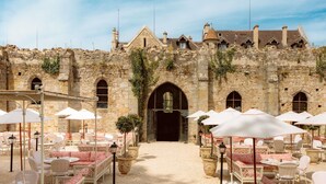 An outdoor café with stone walls and arches, furnished with white tables and chairs, and potted plants.