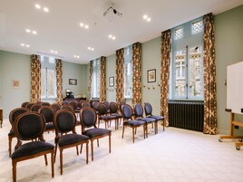 Uma sala de conferências com fileiras de cadeiras, um projetor e grandes janelas com cortinas estampadas.