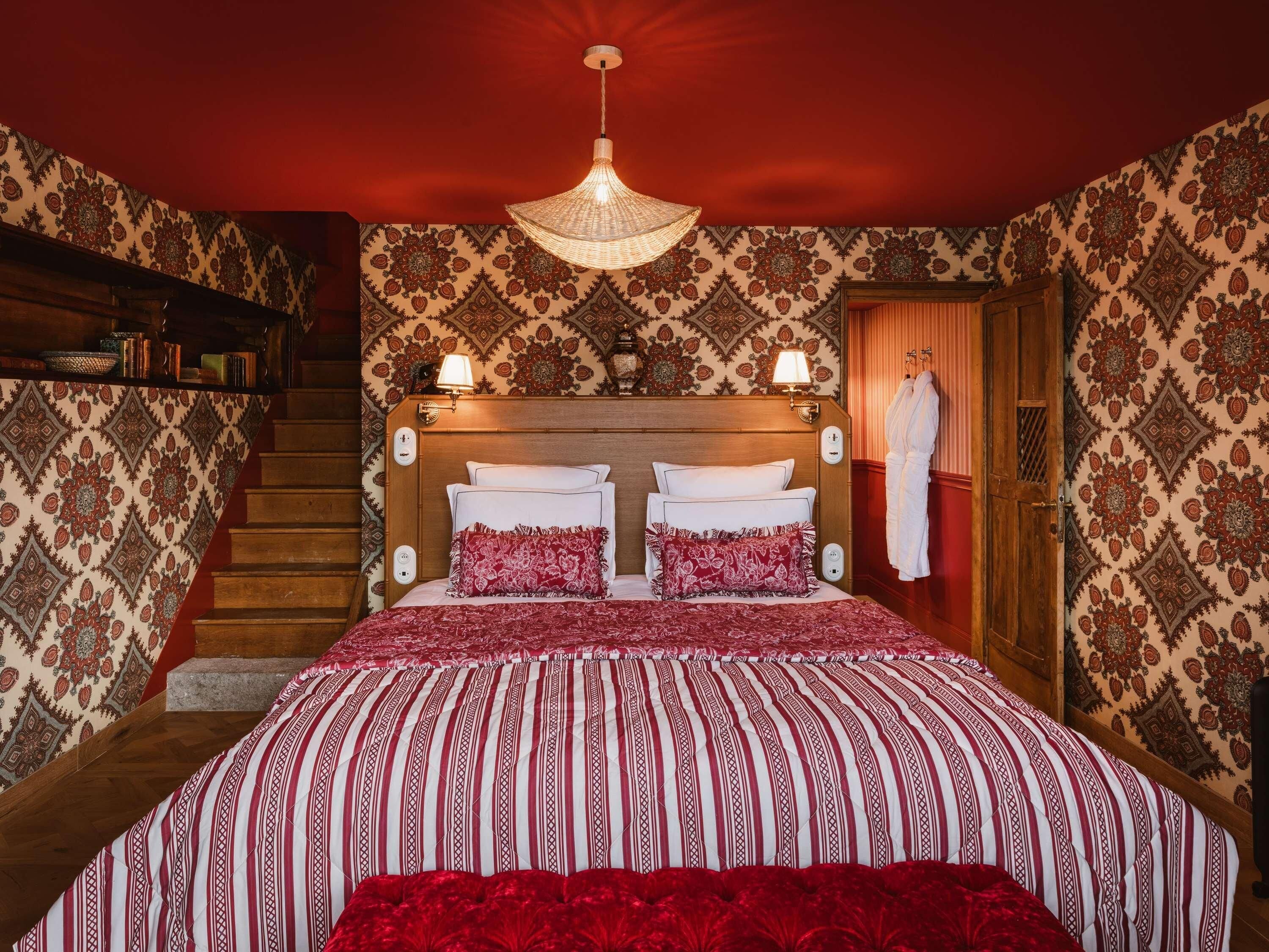 A hotel room with a large bed, a wooden headboard, a chandelier, a staircase, and patterned wallpaper.