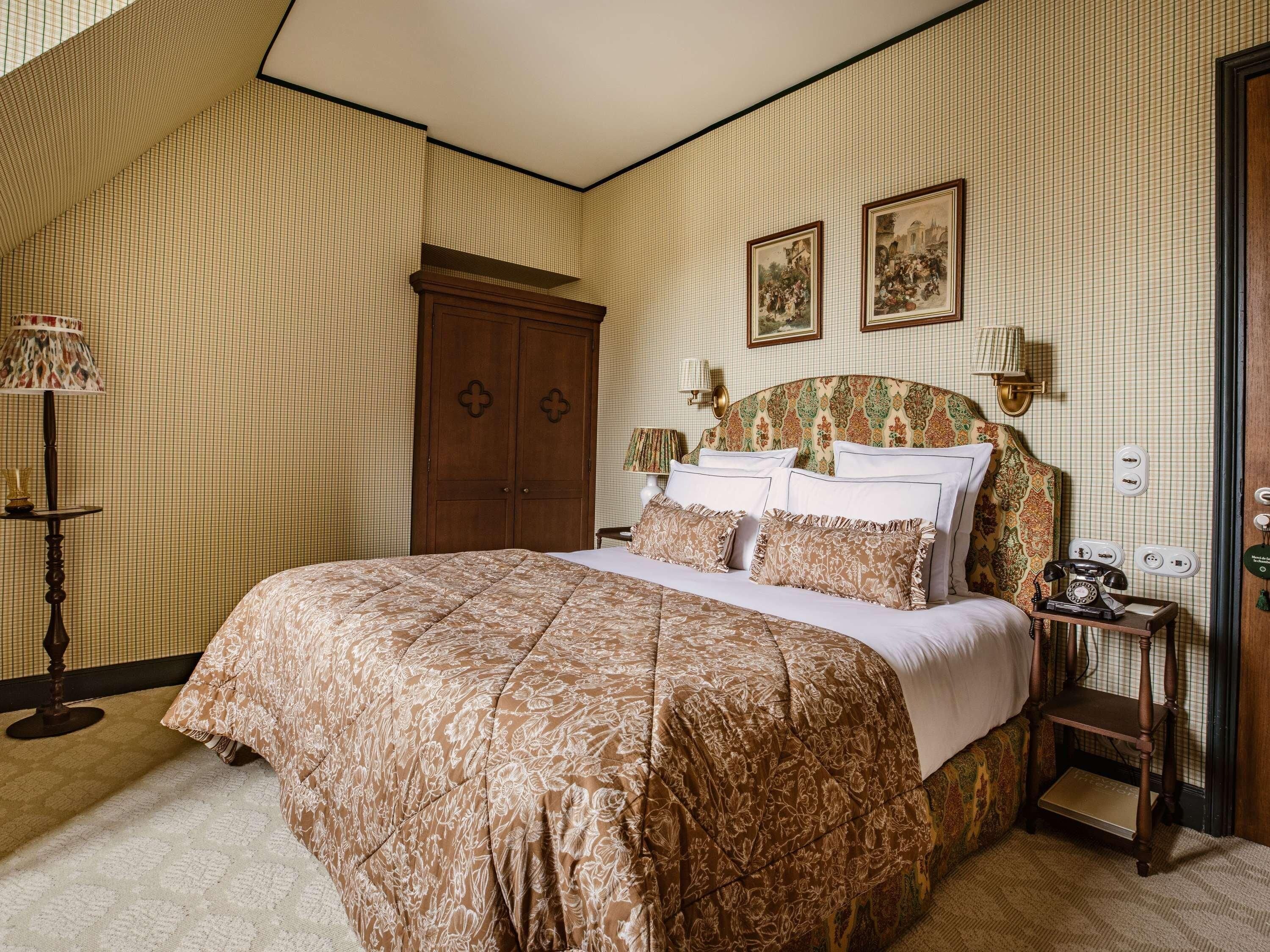 A bedroom with a large bed, a wooden wardrobe, a nightstand with a telephone, and two framed pictures on the wall.