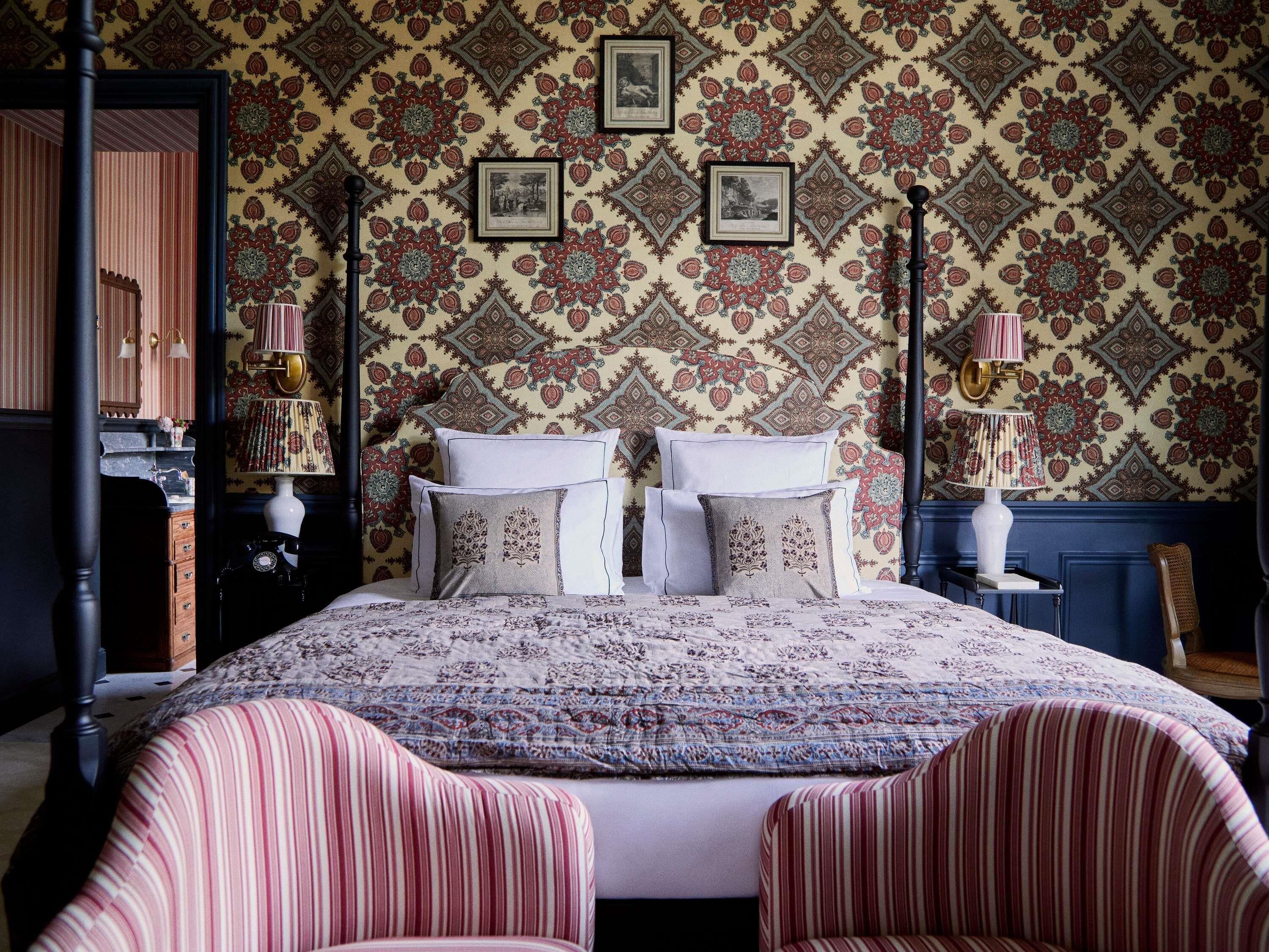 A bedroom with a patterned wallpaper, a bed with pillows, a nightstand, a lamp, and a chair.