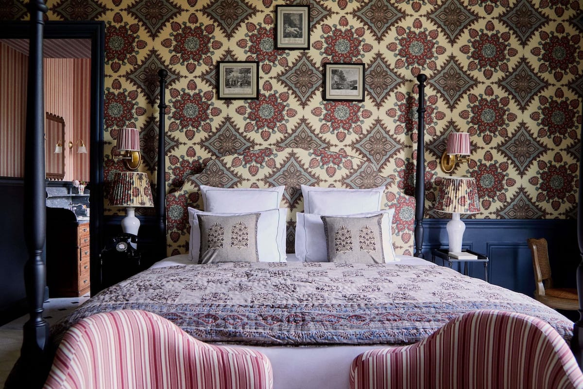 A bedroom with a patterned wallpaper, a bed with pillows, a nightstand, a lamp, and a chair.