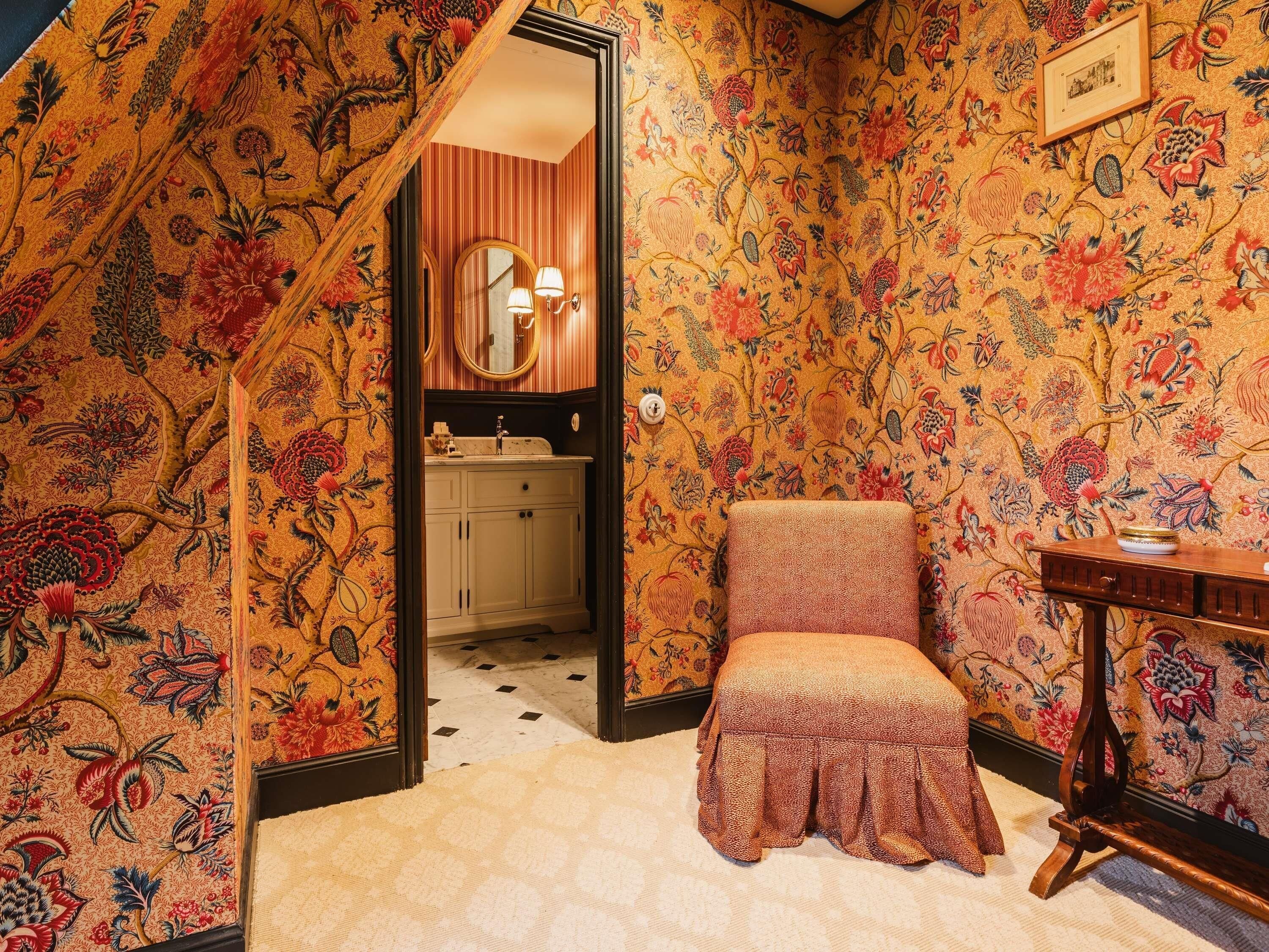 A room with a patterned wallpaper, a wooden chair, a small table, a mirror, and a sink.
