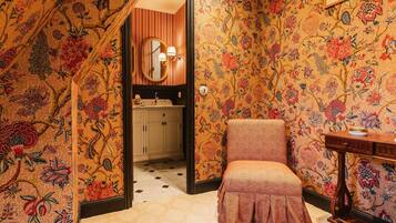 A room with a patterned wallpaper, a wooden chair, a small table, a mirror, and a sink.