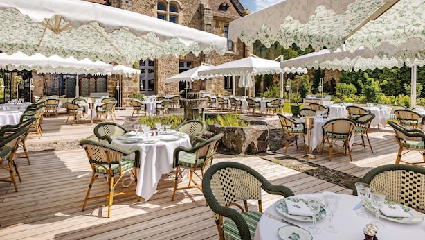 Un área de comedor al aire libre con sillas y mesas de mimbre, paredes de piedra y un edificio grande al fondo.