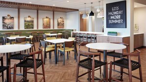 A modern restaurant with wooden floors, round tables, and chairs. There are paintings on the walls and a sign that says "LESS MONDAY MORE SUMMER". - Fairfield by Marriott Inn & Suites Morrow (Morrow)