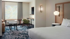 A hotel room with a bed, a desk, a chair, a small table, a lamp, and a large window with curtains. - Fairfield by Marriott Inn & Suites Morrow (Morrow)