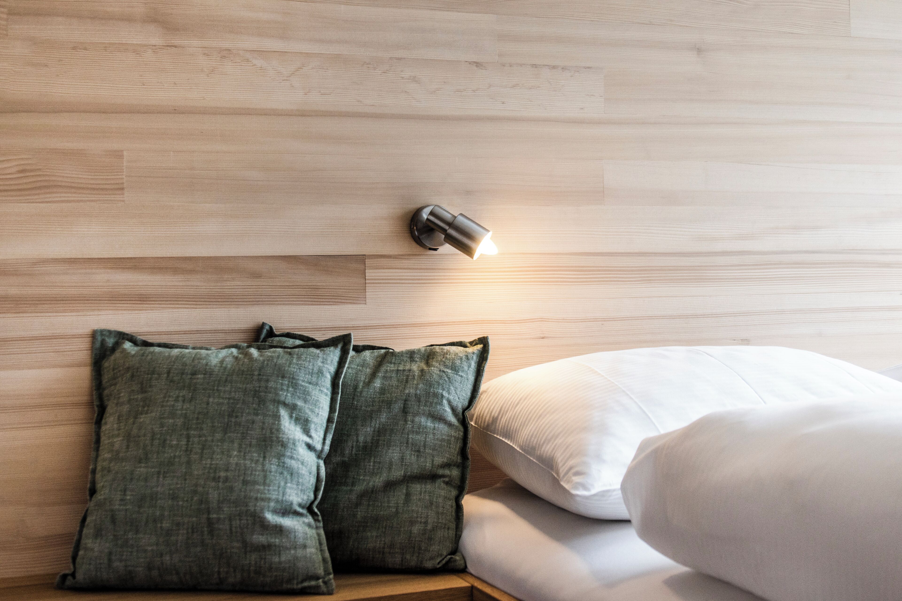 Ein ordentlich bezogenes Bett mit zwei Kissen, ein Kopfteil mit einer Wandleuchte und ein Holzboden.