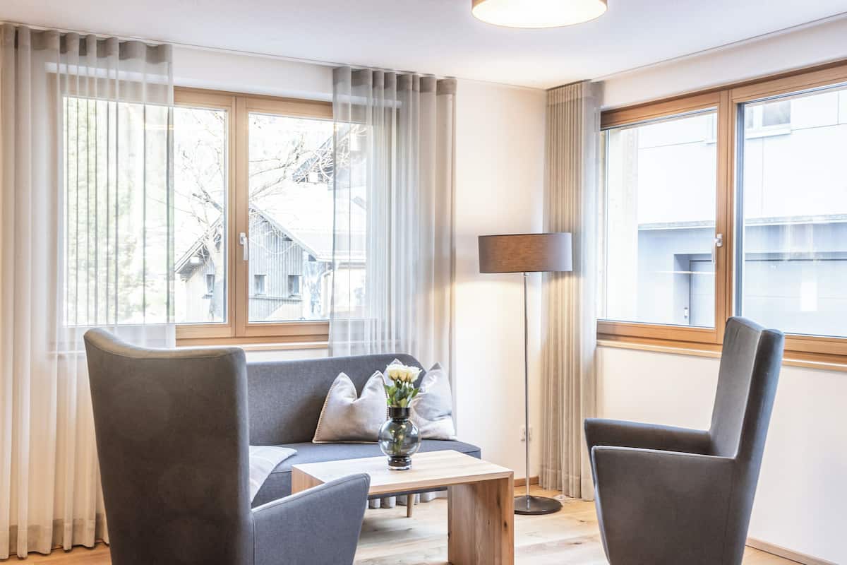 A modern living room with a grey sofa, two armchairs, a wooden coffee table, and a floor lamp. Large windows with sheer curtains allow natural light to fill the room.
