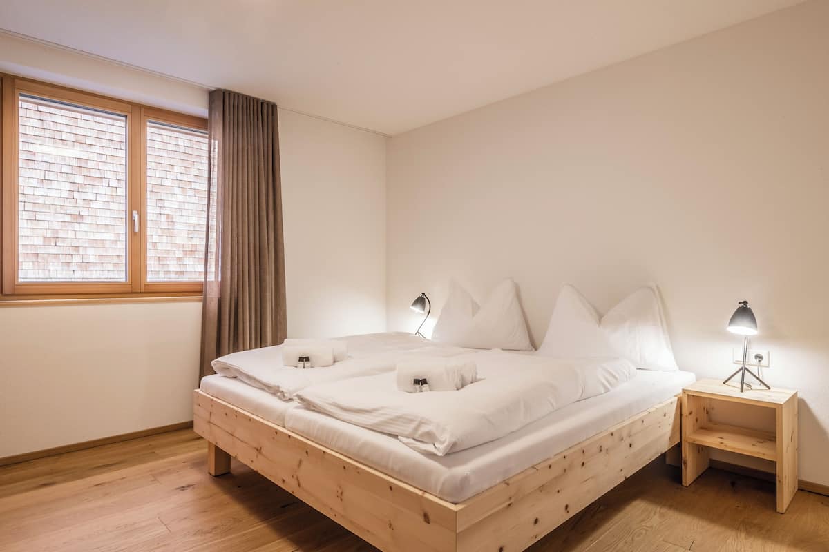 A single-bed room with a wooden frame, white bedding, a nightstand with a lamp, and a window with blinds.
