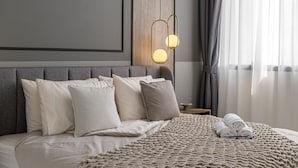 Una cama bien hecha con una manta color beige y almohadas, una mesita de noche con una lámpara, y vistas a una ventana con cortinas.