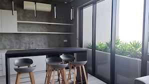 Cocina moderna con una isla grande, taburetes de bar, iluminación colgante y vistas a zonas verdes a través de amplios ventanales.