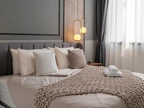 A neatly made bed with a beige blanket and pillows, a bedside table with a lamp, and a window with curtains.