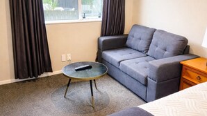 Ein Zimmer mit grauem Sofa, einem kleinen runden Tisch mit Fernbedienung, einem Nachttisch aus Holz und einem Bett mit weißer Decke.