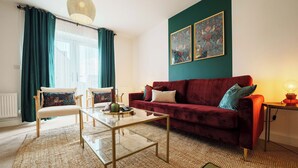 Un salon avec un canapé rouge, deux fauteuils, une table basse en verre et un mur vert orné de deux œuvres d’art encadrées.