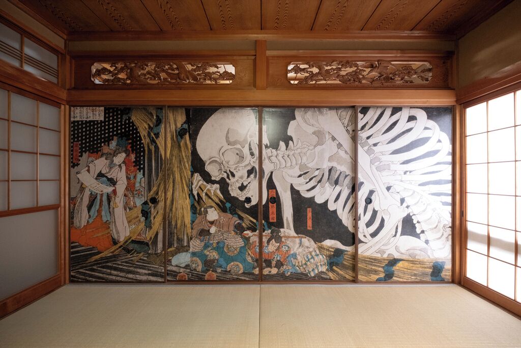 Een traditionele Japanse kamer met tatami vloerbedekking, schuifdeuren en een groot shoji scherm met afbeeldingen van historische taferelen.